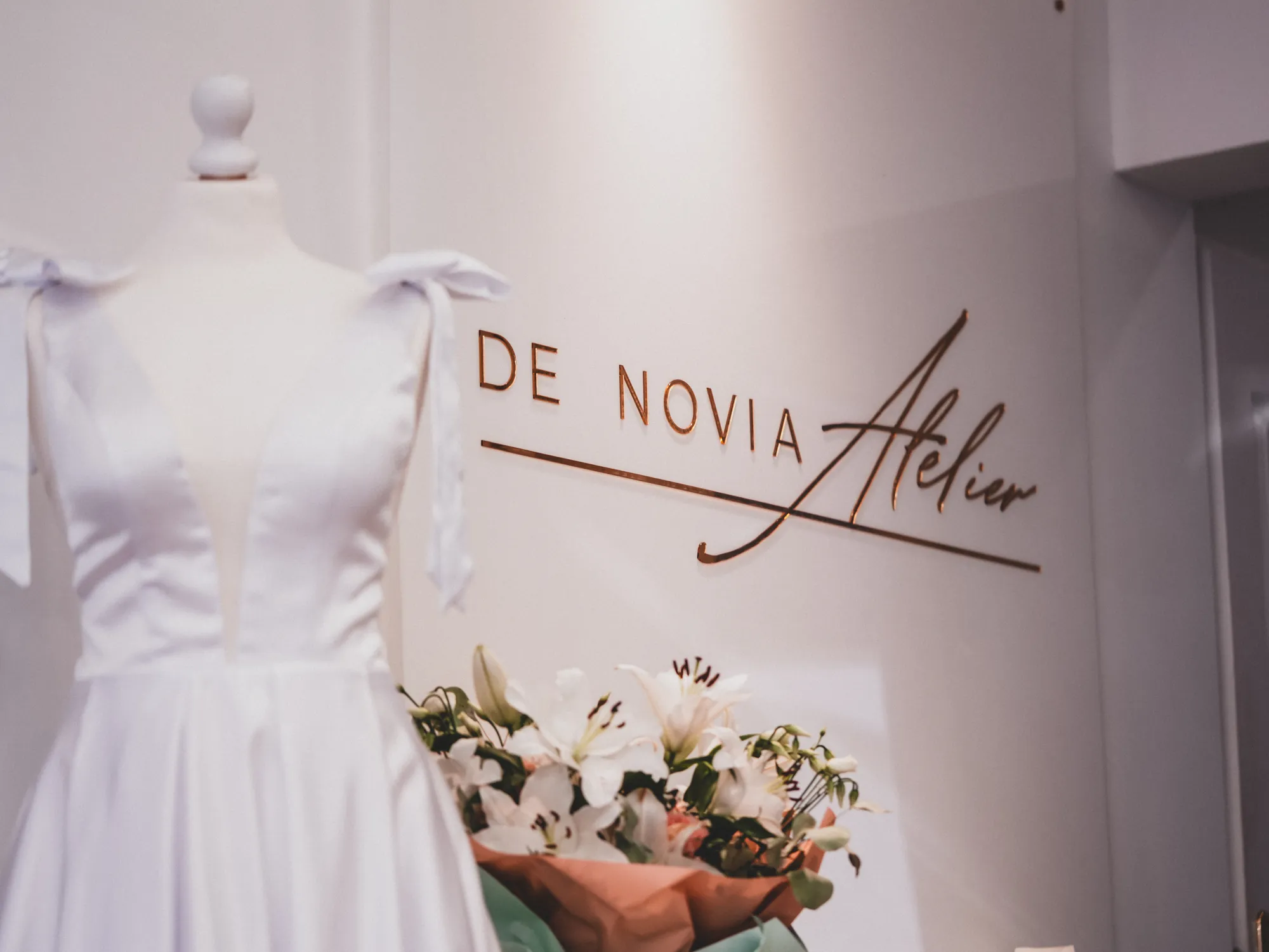 Ściana z logo De Novia Atelier, suknia ślubna na manekinie i lilie